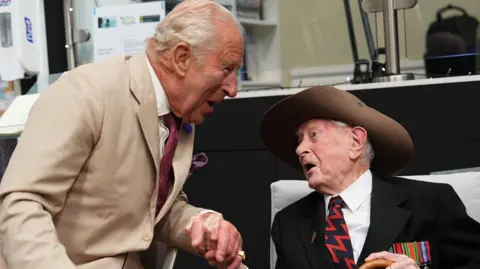 The King, who is wearing a beige suit and red tie, in conversation with James Fenton, who is wearing his slouch hat and has medals on his chest.