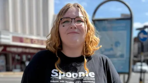 Charlie Lincoln has short wavy hair. She is standing outside and is wearing a black T-shirt which says Spoonie in white writing. She is wearing glasses and is looking directly at the camera and is smiling.
