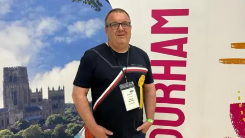 Terry Rooney smiling into the camera with his hands in the front pockets of his trousers and is standing in front of a bored which has a photo of Durham Cathedral on it. He is wearing a black top with a red and white stripe across it. He has short, dark hair and is wearing glasses.