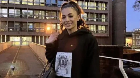 A woman stands outside a grey, brutalist court building at dusk. She is smiling. She has her hair in a high ponytail and holds a handbag at her elbow. She is wearing a black hoodie with a black and white picture of a bulldog on the front.