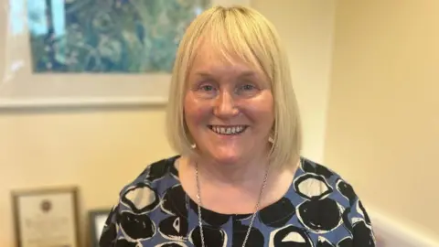 A woman with blonde, shoulder length hair, smiling at the camera. She is photographed against beige walls, from the chest up, wearing a patterned black blue and white top with a silver chain necklace