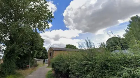 Google Streetview Alderthwaite Farm in Hoyland, Barnsley. A farm track with a hedge on the right, a corn field on the left, blue sky with white clouds, and an old barn at the end of the road