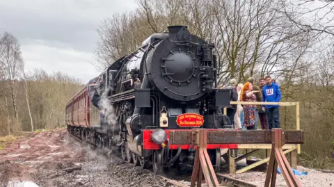 Viewed from in front, a black locomotive is stopped against a makeshift wooden platform. The engine is pulling a series of maroon carriages, and there are a group of people stood on the makeshift platform. 