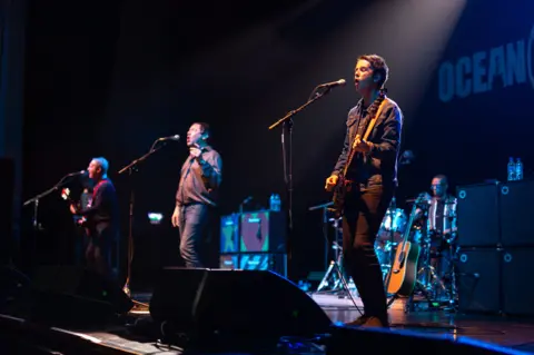 Getty Images Three musicians stand in a line on stage and we look at hen from the right side of the stage. Two have guitars and the middle man is singing. A drummer sits behind them and speakers are stacked up either side of the drum kit.