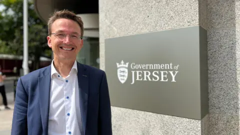 Kirsten Morel is pictured. He is wearing a navy suit with a white shirt underneath and he is smiling. He is standing next to the Government of Jersey sign on a grey building wall.