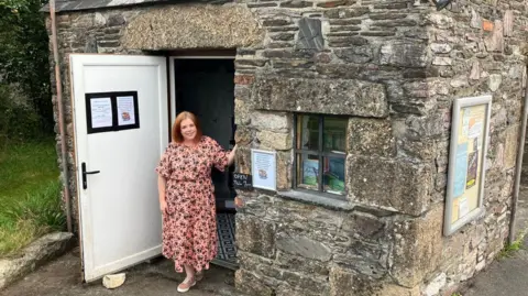 BBC A woman, with a pink dress, standing in the doorway of a small building. The door is open, on the left, and has a poster on it. There is a window on the right and two books on the windowsill.