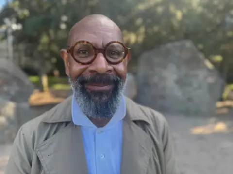 BBC/Simon Atkinson A man with brown rimmed glasses and a beard smiling at the camera while dressed in a light blue shirt and grey jacket. 
