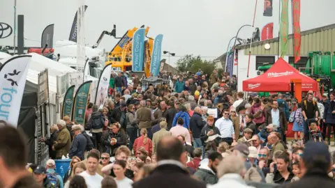 Matt Keeble/PA Wire Hundreds of people at the Royal Cornwall Show which is held in Wadebridge. Stalls with large flags can been seen to either side and large yellow farm machinery in the background. 
