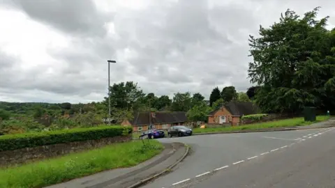 A road junction, with cars parked on the street which is dipping down a hill. There are street lights and houses as well as a pavement and a large tree near the junction.