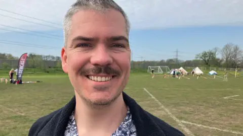A man smiling with grey hair and a black moustache. He is wearing a floral shirt with a jacket over the top. In the background are several tents standing on a mown grass field.