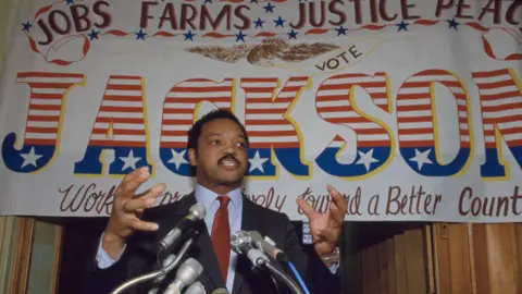 Getty Images Jesse Jackson is standing in front of a sign that reads 'jobs, farms, justice, peace'  'vote Jackson' as he is speaking at one of his campaigns. 