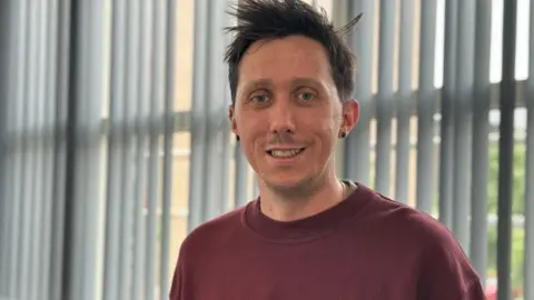 A man with brown spikey hair in front a window. He is smiling and wearing a burgundy T-shirt.