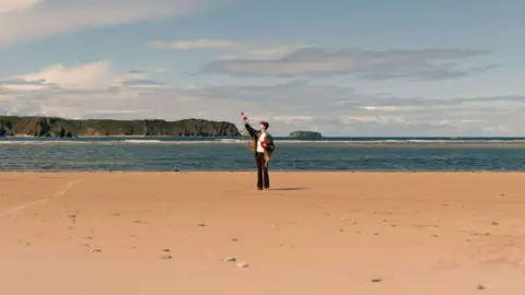 George Houston A man wearing a blue jacket stands on the beach. He is far away from the camera. It is a sunny day, and the sea is visible behind the man.