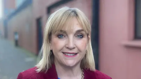 Celine McStravick has long blond hair and blue eyes. She is smiling at the camera. She is wearing a blue jumper and a maroon blazer. Behind her is a red wall. 