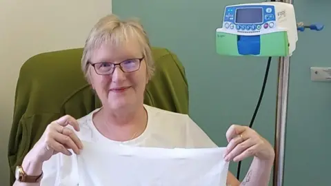 A woman, Christine Campbell, sitting in a chair in a clinical setting. She is holding a sheet with information about ovarian cancer and there is medical equipment around her. she is wearing a white top and dark trousers and is wearing glasses. She is smiling.