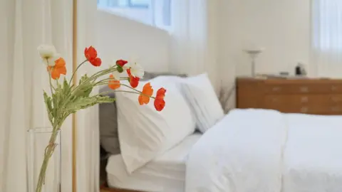 Getty Images A vase of orange and white flowers in a bright bedroom with a neatly made white bed.