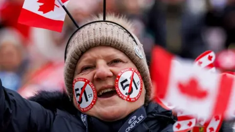 A person in a beige woollen hat and navy jacket and with stickers on the face holds a Canadian flag