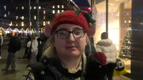 Ms Keene is wearing a red elf bauble hat and a black bodywarmer and top. She has black glasses.