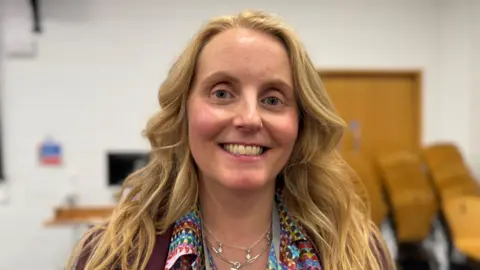 Sharon Edwards / BBC A head and shoulders shot of council leader Naomi Tweddle smiling at the camera. She has long, blonde curled hair and is wearing a colourful, patterned shirt and a burgundy blazer. 