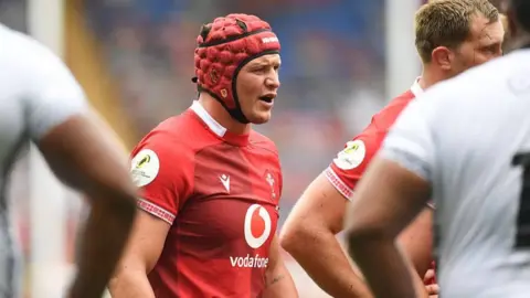 Getty Images James Botham in a Wales shirt. He wears a red protective cap, and stands with other players on the pitch. 