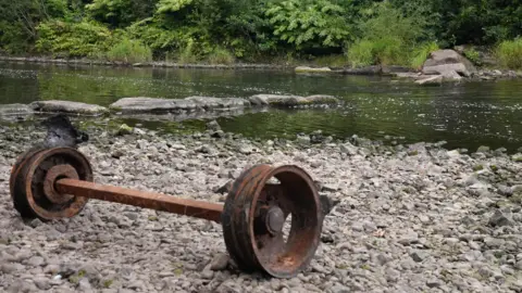 A rusted set of wheels connected by an axle sits on the side of the river on some small stones. The river can be seen behind it with a row of rocks leading into the centre of the water with a row of trees behind.