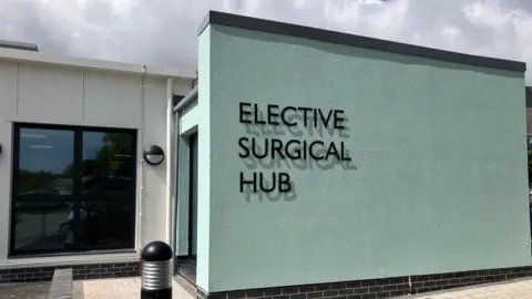 The exterior of St Austell's Surgical Hub. The building is mint green and has the words 'Elective Surgical Hub' on the front in large black letters. To the left is a large window with a single door to the right. 