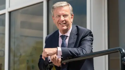 David Webster, a man wearing a dark suit. He is leaning against a metal barrier. In the background is a building with large windows. He is looking at the camera and smiling.
