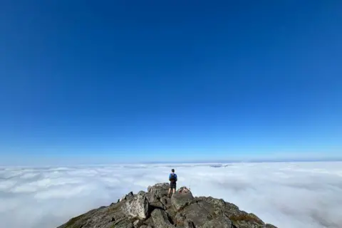 Ashley McLean Man at peak of Schiehallion, above clouds, with blue sky in background