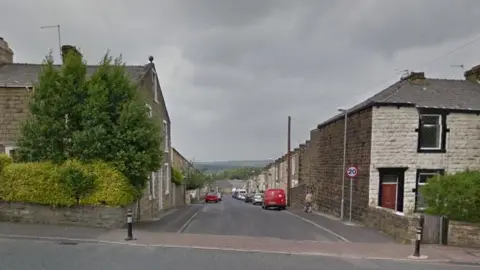 Google streetview looking down Craven Street, which is lined with dark and white brick terrace houses and parked cars. Pennine hills can be seen in the backdrop.
