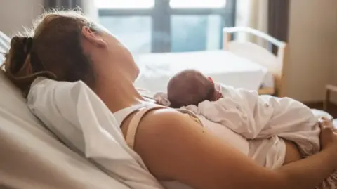A woman is cradling a baby on her chest in a hospital ward. Both are looking away from the camera and wearing white