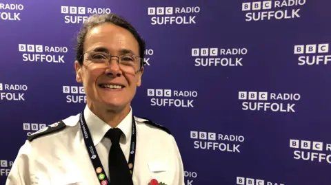 Vikki Irwin/BBC Rachel Kearton, wearing a police uniform and glasses, standing in front of a purple BBC Radio Suffolk backdrop. 