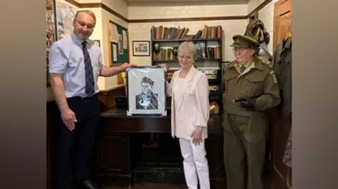 Dad's Army Museum A man wears a blue shirt, tie and black trousers and is holding a black and white portrait of Ian Lavender, which is being held on the other side by Michele Hardy. She is wearing a cream jacket and shoes, and a white top and trousers. They are standing next to a life-size model of a Dad's Army character.