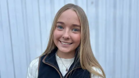 Georgie Nairn with long blonder hair and smiling to the camera, wearing a blue fleece gilet over a cream top