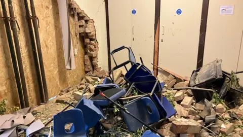 Shariqua Ahmed/BBC Wrecked meeting room- with bricks and debris on the floor- blue chairs and table damaged and thrown in the rubble
