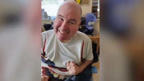 Family photo A man with a bald head is sat and smiling at the camera. He is wearing a white t-shirt with blue and red patterns on it.
