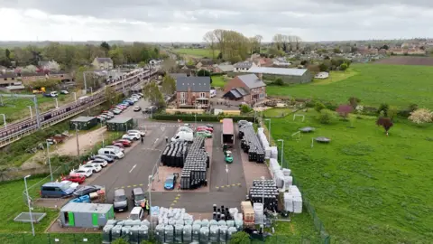 Qays Najm/BBC Aerial view of thousands of black bins in a car park, with a lorry in the background and a forklift truck carrying bins around. There are cars in the car park which has a rail line to the left and fields to the right.