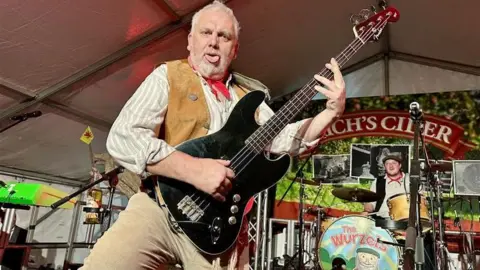 Bassist holding bass up to the camera with his tongue out and the drummer in the background