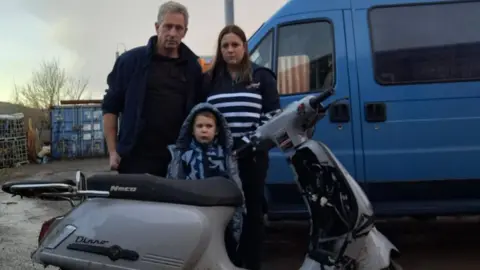 Becca, John and Joseph are standing behind the Neco Dinno scooter which was stolen last week. It has been damaged and the family look unhappy. Behind them is a blue van.