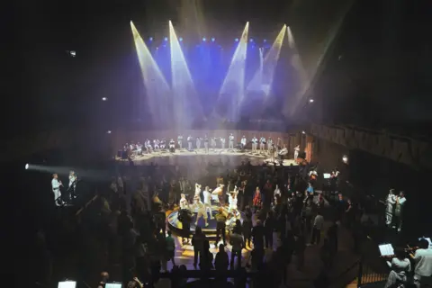 A performance by Paraorchestra at BBC Proms in Bristol in 2024. The stage is lit with blue lights and spotlights. Silhouettes of the orchestra members can be seen. 