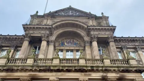 An external shot of the upper floor Birmingham Council House, which is a sandstone building with moulded columns and a balcony. Statues of lions line the balcony on plinths and a central arch sits behind it, featuring a mosaic. A pale blue sky can be seen above.