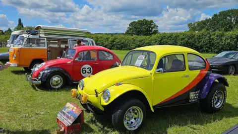 Two brightly coloured modified VW Beetle cars in a field, next to a VW Camper van. 