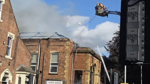 Paul Scotson Firefighters stand on a cherry picker over a smoking building with a damaged roof