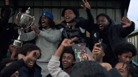 MoFilmedit A silver metal cup is waved in the air by a fan surrounded by other fans on what appears to be part of a football terrace, with metal sheets at the back behind them. The fan with the cup has a blue cap on backwards, a grey top and grey trousers while the other fans have puffy black jackets, some hold phones aloft as if recording the scene.