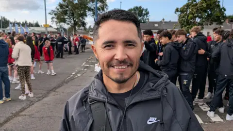 Head coach Hugo Chaparro is wearing a team tracksuit and standing in a car park.
