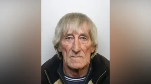 A mug shot of a man with long grey hair, blue eyes, wearing a navy jumper with white stripes and a jacket.