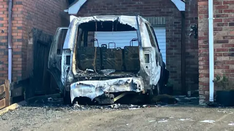 BBC A burned out jeep sits in the drive way of a house. it is seen from behind looking into the boot and is completely destroyed by fire. A garage stands behind it while the driveway is covered in detritus 