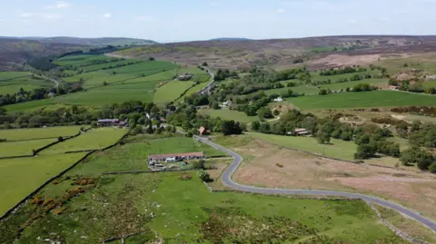 The remote village of Commondale - in the North York Moors National Park - is seen from above. The fields - divided by stone walls - are various shades of green. A road snakes through the fields and up towards the moors, with hills in the distance.