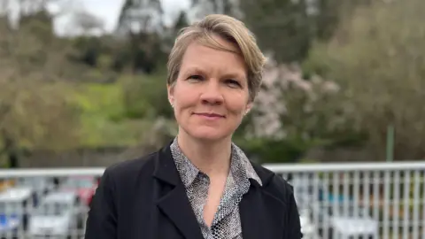 Deputy Sasha Kazantseva-Miller - A woman with short blonde hair, wearing a black jacket and a brown and white shirt. 