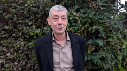 An image of a man wearing a purple shirt and black blazer, standing in front of a hedge. He has grey hair and is wearing a silver chain.