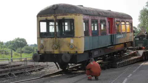 North Dorset Railway A rail carriage with scruffy paintwork is rolled from a low-loader on to a railway track